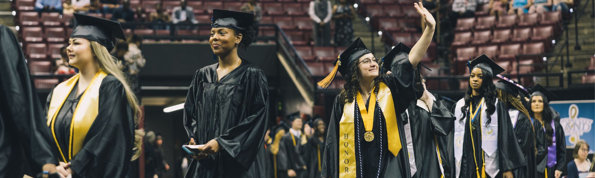 Honors student at graduation smiling and waving up at the crowd.